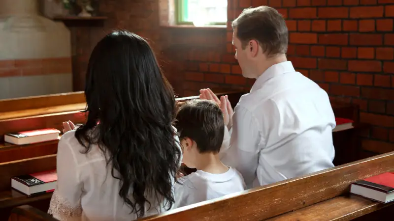 A family of three sits in wooden pews, hands clasped in prayer, surrounded by a brick church interior and floral decorations