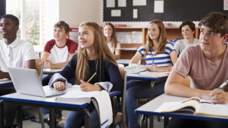 A child wearing headphones sits at a desk with educational materials, while an adult gestures and teaches using a laptop